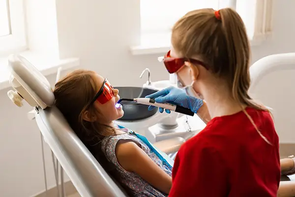 A dentist treating a young girl by applying a dental sealant with a curing light, with both wearing protective glasses in a brightly lit dental office.