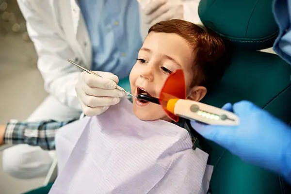 A child receiving a dental sealant application using a curing light, assisted by a dentist wearing gloves, in a modern dental clinic.