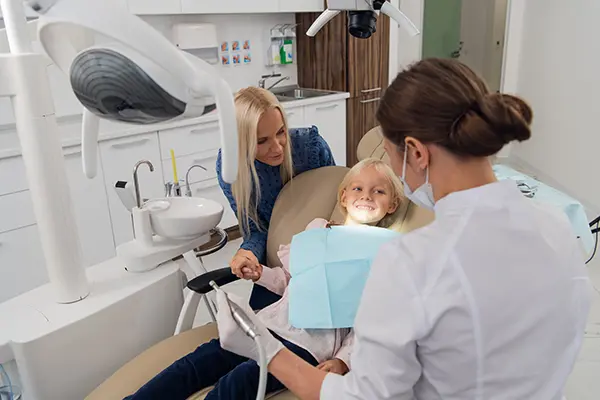 A dentist examining a young girl's teeth while her mother offers support in a modern dental clinic.