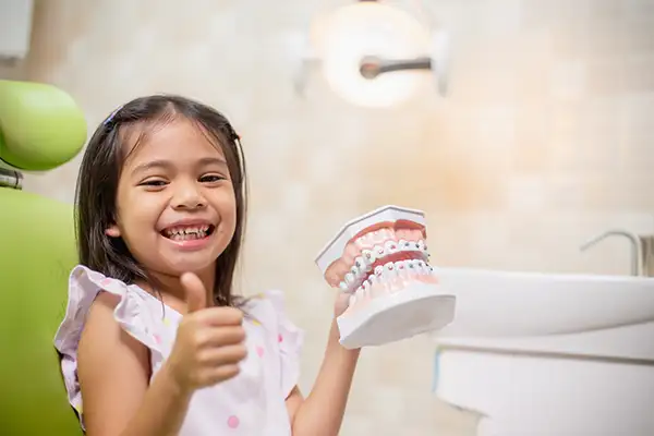 Smiling young girl holding a dental model and giving a thumbs-up at a dental clinic, promoting early orthodontic care.