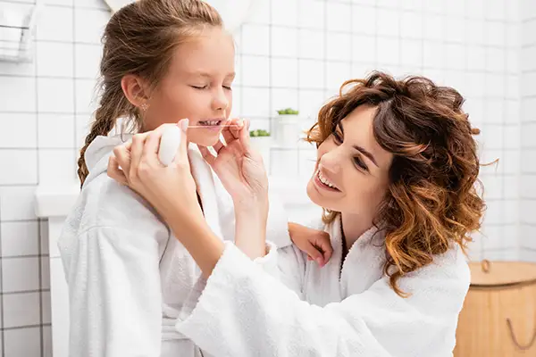 Mother assisting her young daughter with flossing her teeth in a bathroom, encouraging early oral hygiene habits.