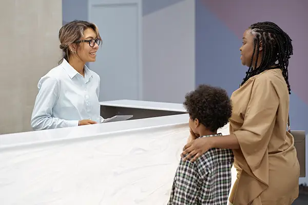 A friendly patient care coordinator greets a mother at the front desk, checking in before her child's dental visit.