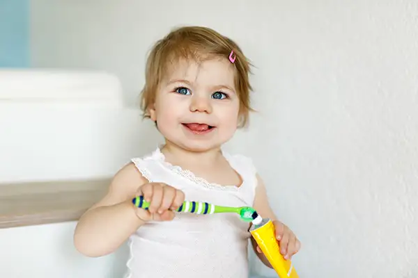 Happy toddler holding a toothbrush and toothpaste, promoting the importance of early dental care for children.