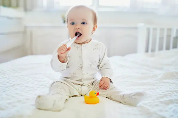 Baby sitting on a bed and chewing on a toothbrush, showing the beginning stages of oral hygiene habits for infants.