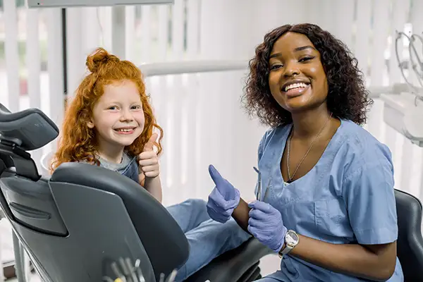 A smiling young girl and her dentist giving thumbs up from the dental chair, in honor of a great dental checkup.