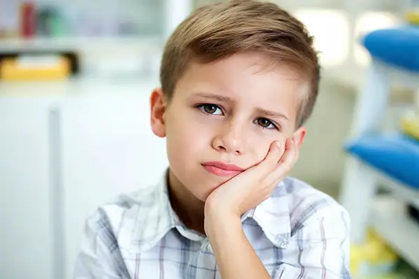 Young boy sitting in a dental clinic, holding his cheek and looking distressed due to tooth pain, requiring emergency dental care for kids.