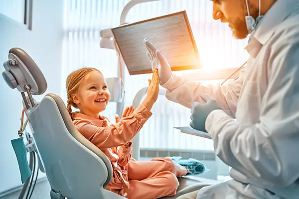 A smiling young patient seated in a dental chair high fives her pediatric dentist, demonstrating the importance of skill and connection when practicing dentistry for kids.