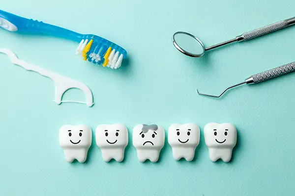 Dental tools, a toothbrush, and tooth floss surrounding smiling tooth models with one showing a sad expression due to its cavity.